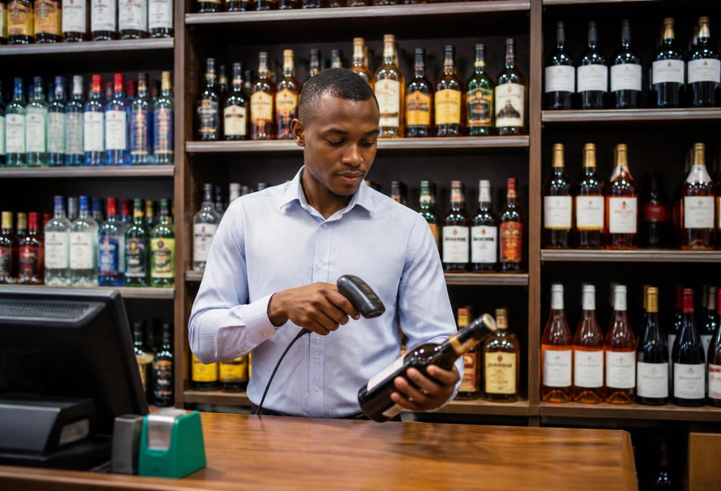 Liquor store cashier in Tanzania scanning a bottle at a StellarPOS point of sale terminal with shelves of beer, wine and spirits behind